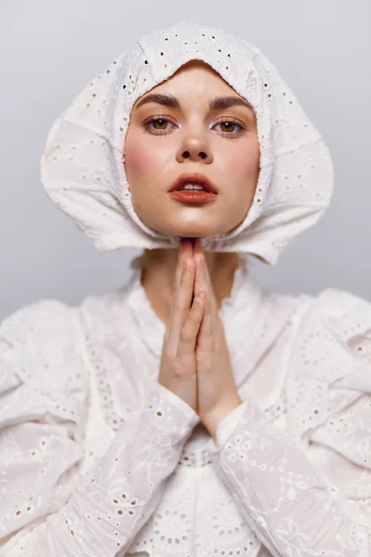 A young woman with a serene expression wears a vintage white outfit and a bonnet, showcasing a delicate and timeless style against a neutral backdrop