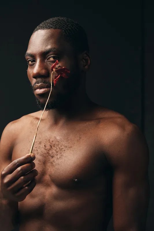 Creative conceptual portrait of African man with dark skin holding dried flower near face in studio. Artistic visual style with bare chest and thoughtful expression on black background.