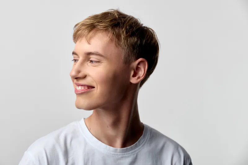 Young man with a bright smile turns slightly to the side, wearing a plain white tee, casual and confident, studio light highlights his cheerful expression for lifestyle and fashion themes