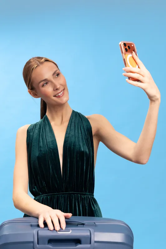 Young woman in elegant dress taking selfie with smartphone while holding suitcase on blue background. Concept of travel, vacation, lifestyle, and modern technology use for communication.