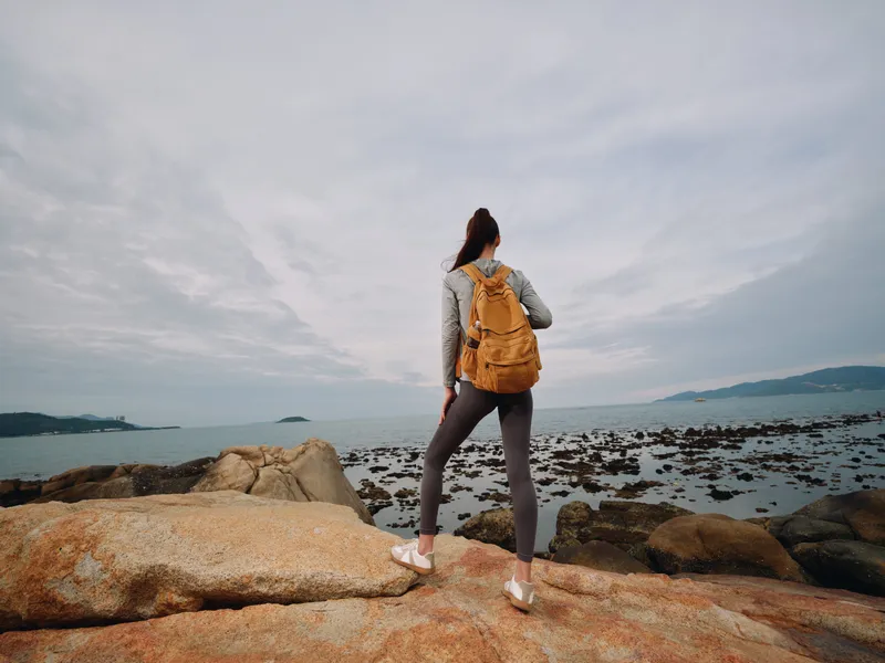 Young woman enjoying nature on rocky coastline with backpack, wearing sporty outfit, peaceful mood, outdoor adventure, sea view, travel concept