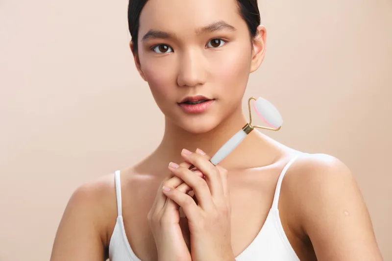 asian woman gently uses a facial roller in a serene skincare setting, conveying calm confidence and self care. soft lighting highlights smooth skin and a focused, tranquil mood.