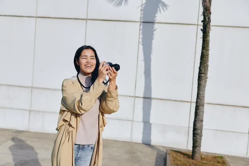 Smiling woman with camera capturing a sunny outdoor moment beside a white wall and palm trees, casual fashion and candid photography, lifestyle shot highlighting creativity and exploration