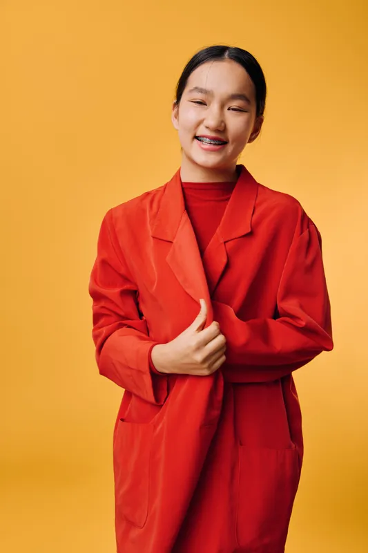 Young smiling woman in red coat standing against orange background with eyes closed and happy expression. Casual style, female model posing with hands crossed in cozy outfit.
