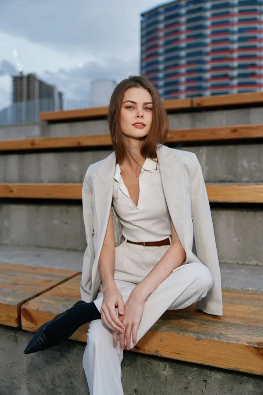 confident woman sitting outdoors in beige business outfit with light jacket, urban background, casual fashion style, modern businesswoman portrait