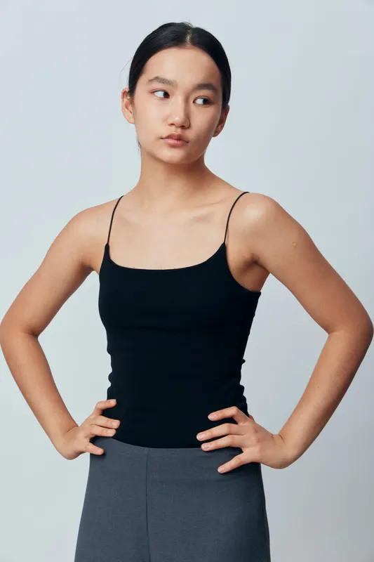 Young woman in black tank top and gray pants poses with hands on hips against plain background. Casual style portrait showing confident and relaxed mood indoors in natural light.