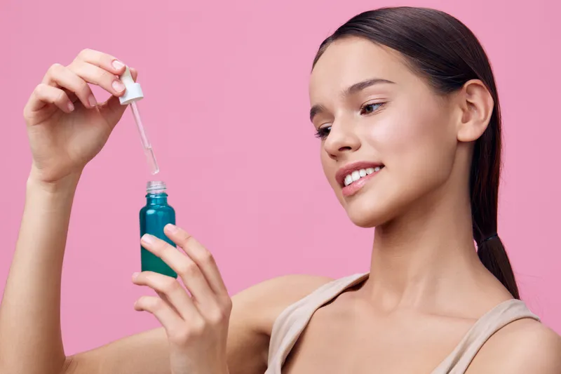 Young woman applying skincare serum from a dropper bottle, showcasing healthy skin and a joyful expression against a pink background
