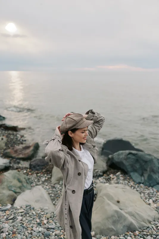 Young woman in beige coat and hat stands on rocky beach near calm sea at sunset. She adjusts her hat while enjoying peaceful nature and fresh air by the water, concept of relaxation and freedom.