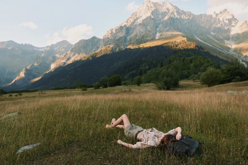 A young woman lies on a sunlit meadow with rugged mountains in the background, resting beside a backpack, conveying relaxation and a sense of outdoor freedom.
