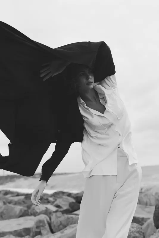 Fashion movement with cape billowing as wind lifts fabric over a beach setting, elegant white outfit, dramatic monochrome mood and dynamic pose.