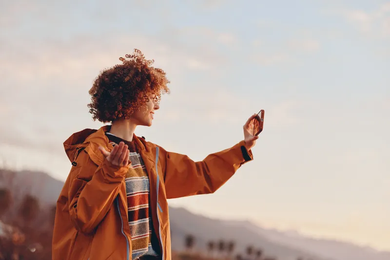 Woman lifestyle outdoors in autumn coat and scarf, sunrise sunlight, smiling while stretching hands, soft breeze, scenic mountains background, relaxed confident mood