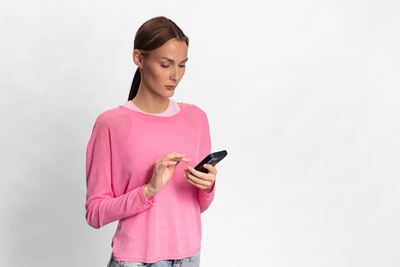 Young woman in pink sweater stands against white background, focused on smartphone in hands. Modern casual style, technology use, communication and digital lifestyle concept.