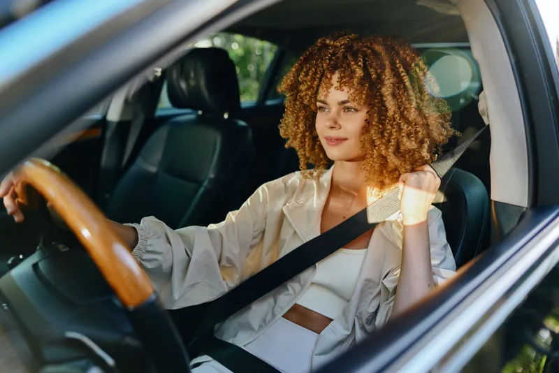 Curly woman wearing seatbelt driving car in bright natural light, displaying calm focus and safety awareness during daytime on a sunny day.