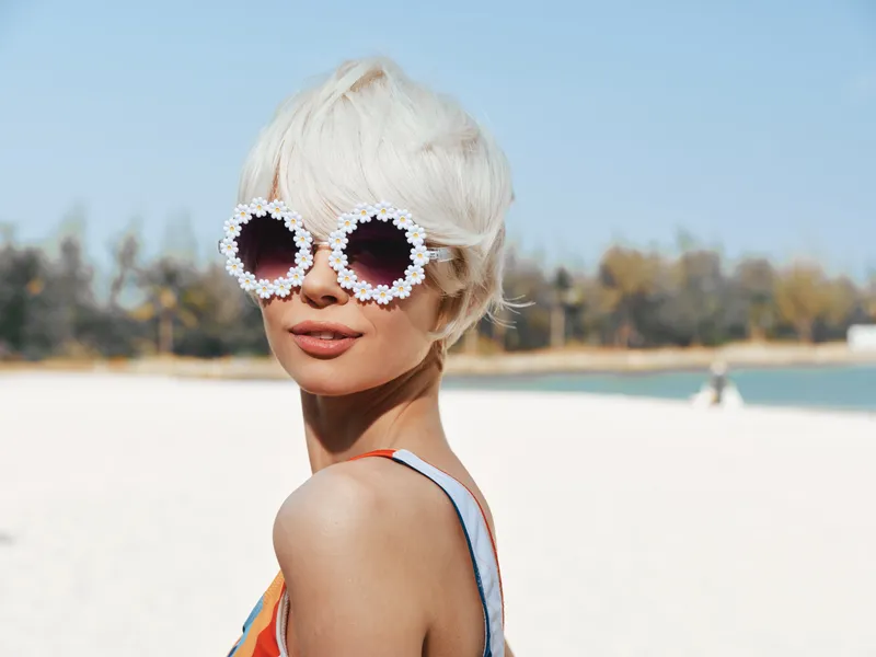 Stylish beachgoer with oversized floral sunglasses and short platinum hair poses on a bright sunny shore, showcasing playful summer style and confident, carefree charm.