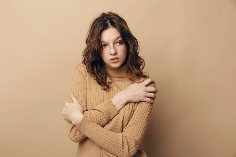 Young woman with curly hair wearing beige sweater hugs herself, looking away with thoughtful expression. Neutral background creates calm and introspective mood.