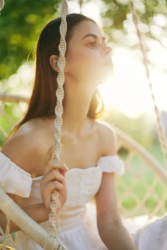 Young woman in a white dress on a swing, enjoying the sunlight, showcasing beauty and serenity in a natural outdoor setting