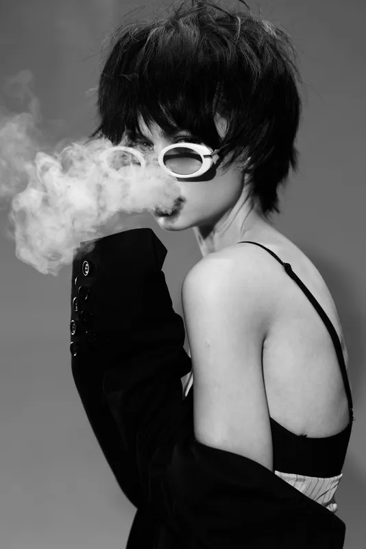 Stylish woman with short dark hair wearing white sunglasses exhaling smoke in black and white artistic portrait.