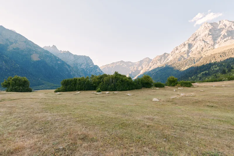 Meadow mountains valley grassland bushes landscape nature sky panoramic alpine meadow with dry grass and scattered shrubs in an open valley beneath rugged mountain peaks and clear sky.