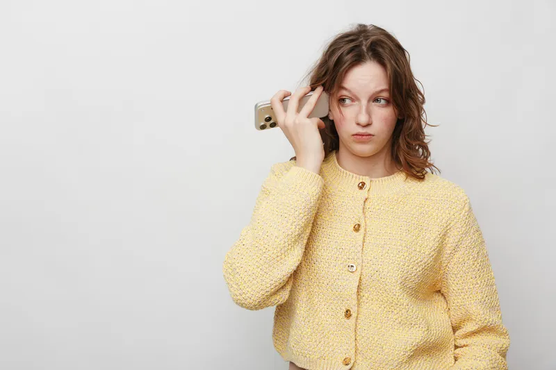 Young woman in yellow sweater holds smartphone near ear, looking away with thoughtful expression. Isolated on white background. Concept of communication, technology, and contemplation.