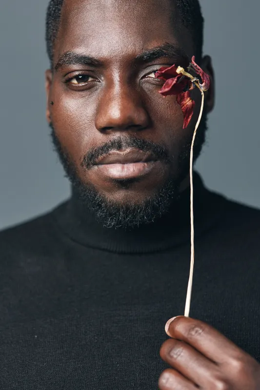 African man with dark skin holding dried flower near face in creative conceptual visual style. Portrait with serious expression and artistic mood on neutral background in studio.