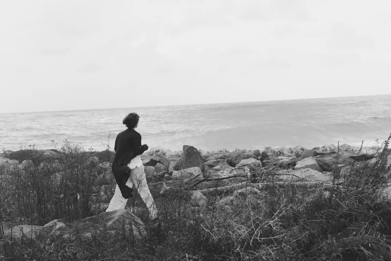 Man walking along a rocky shore toward the sea in a monochrome scene, capturing solitude, rugged coast and distant horizon, atmospheric landscape photography