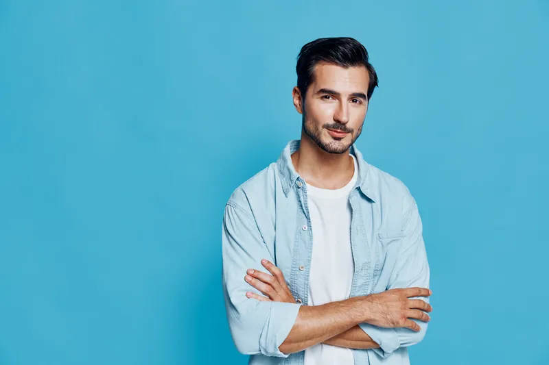 Confident young man with stylish hair wearing a light blue shirt smiles while crossing his arms, posing against a solid bright blue background. Lifestyle concept.