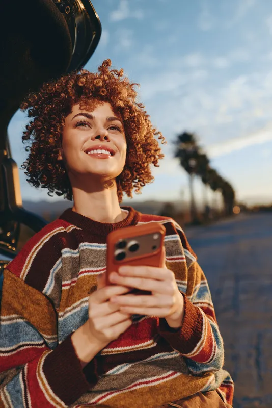 Woman lifestyle outdoors, smiling with curly hair, holding a smartphone near a car, wearing a colorful sweater, golden sunset light, relaxed candid travel moment, natural tones