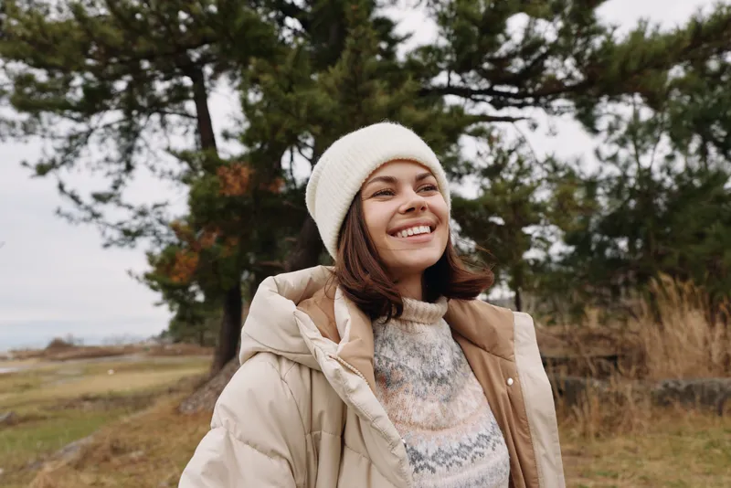 Woman in a warm beige coat and knit sweater stands outdoors among trees, smiling under a soft beanie, enjoying a crisp day in a wide park setting with autumn light and open scenery.