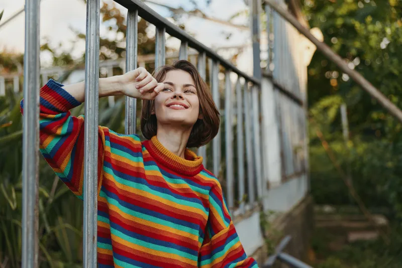 Woman in a striped sweater leaning against a greenhouse metal rail, smiling and touching her face, short hair, casual outfit in a sunlit outdoor garden lifestyle moment.