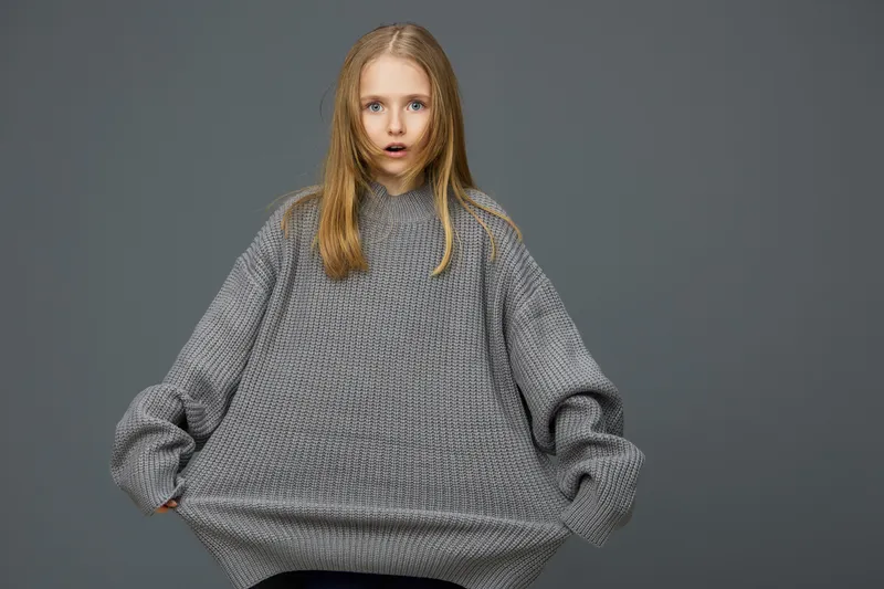 A surprised young girl wearing an oversized grey sweater against a muted background, showcasing a sense of curiosity and playfulness in her expression