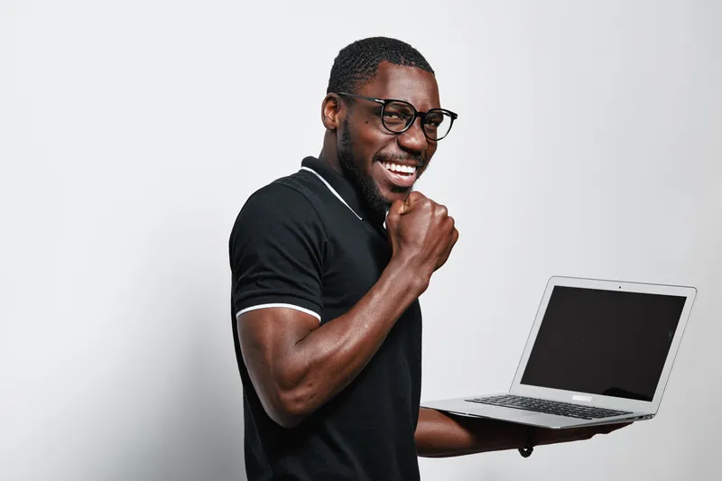 African American man with dark skin holding laptop and smiling in casual black polo shirt and glasses. Technology, communication and digital device for work or study on white background.