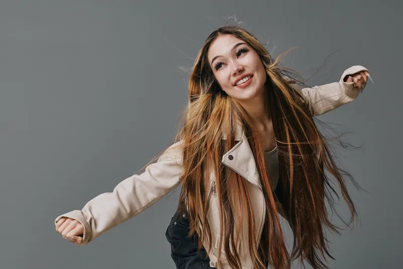 Young woman with long hair wearing beige jacket smiling while posing with arms outstretched on gray background in studio. Happy person, motion and carefree mood with casual style and emotion.