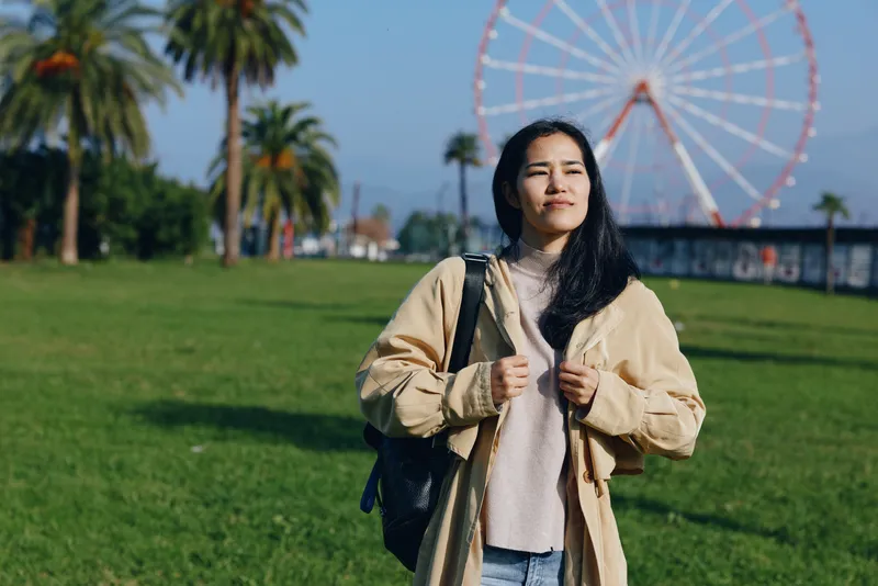 Woman in a beige jacket stands in a sunny park with a large ferris wheel behind, carrying a backpack, enjoying outdoor scenery and fresh air.