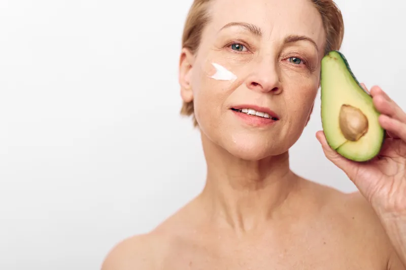 Mature woman with clear skin applies natural cream on cheek while holding fresh avocado half. Concept of organic skincare, healthy lifestyle, natural beauty, and anti aging care.