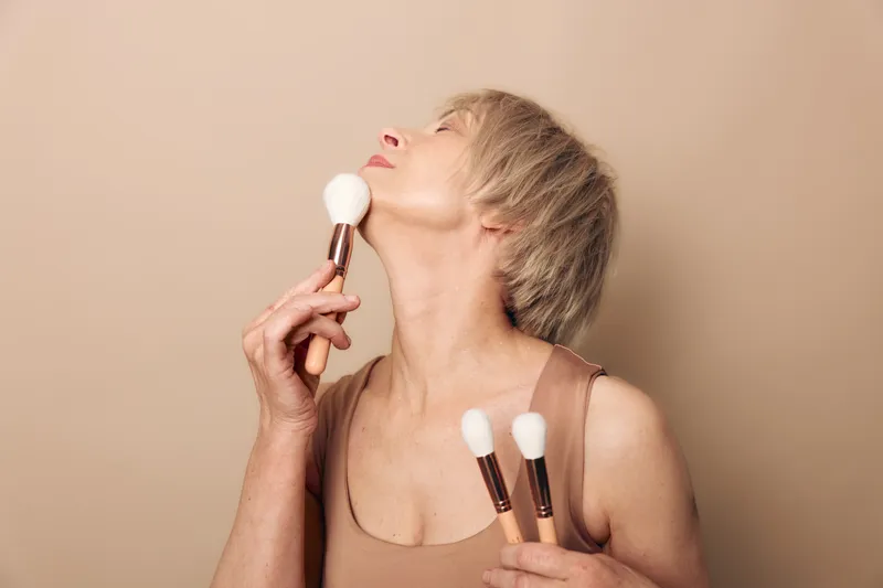 Mature woman with short blonde hair applying powder makeup with brushes on beige background. Concept of beauty, skincare, self-care, and graceful aging in natural light.
