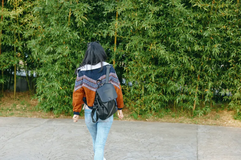 Backpack, walking, woman, outdoors, greenery, sidewalk, street, fashion, sweater, jeans with a lone shopper strolling away along a paved path beside lush hedges and calm urban scenery