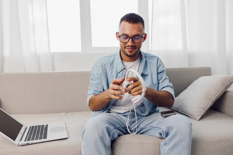 Smiling man adjusting headphones while using laptop in bright modern living room, showcasing technology and comfort in daily life.