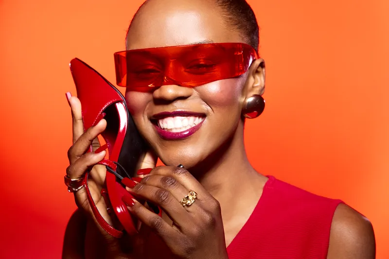 Happy African woman with dark skin smiles wearing red dress and sunglasses holding red high heel shoe near face on orange background. Concept of fashion, style, beauty, and confidence.