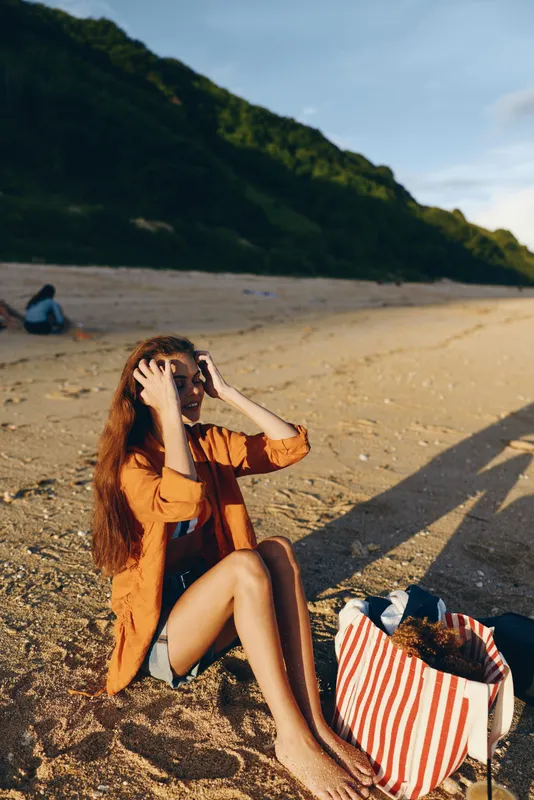Woman beach sand sunlight summer seated and brushing hair, relaxed smile, striped bag beside her, warm golden light, soft breeze, green mountains in background, coastal shore