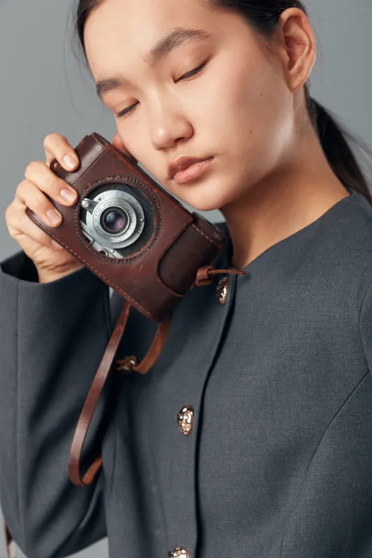 Young woman holding vintage camera close to face with closed eyes wearing dark coat, portrait of photographer with retro equipment and calm expression in studio with neutral background.