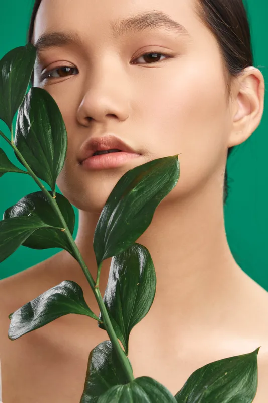 asian woman is framed by glossy green leaves in a vivid studio portrait, presenting natural beauty and poised confidence that communicates a calm, modern mood.