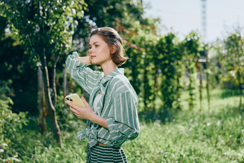 Young woman outdoors holding a phone, standing among trees with bright sunlight, wearing a striped shirt, and gazing thoughtfully.