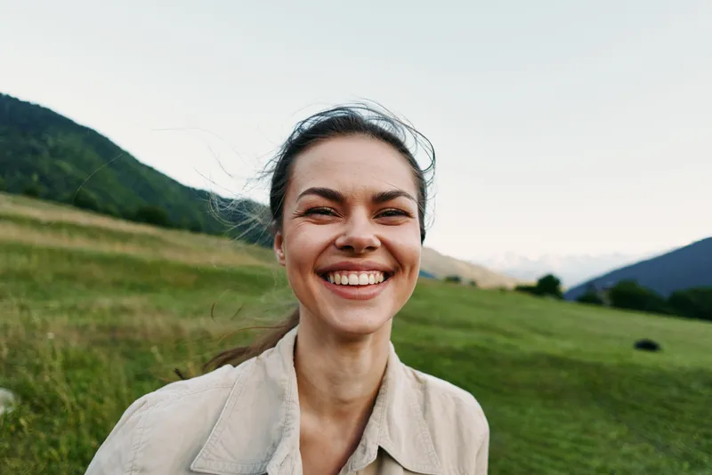 A young woman in a beige jacket smiles widely in a bright outdoor field with distant mountains, capturing a candid, carefree lifestyle moment for a sunny day shoot.