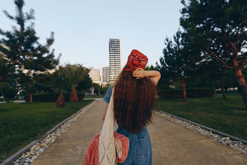 lifestyle woman in modern boho-western denim street style with long hair and red bandana walking urban park at sunset in film color tone