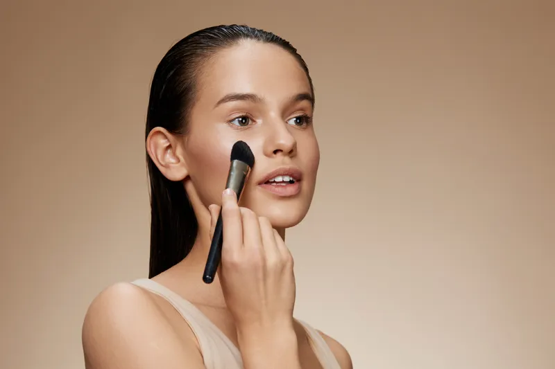 Young model applying makeup with a brush, showcasing natural beauty and confidence, against a soft beige background for beauty and skincare concepts
