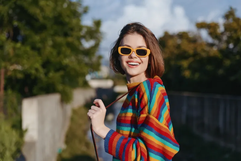 Woman, smiling, striped sweater, yellow sunglasses, young adult, short hair, outdoor, walking — 20s female with bright rainbow knit, turning and laughing in sunny urban park for lifestyle travel.