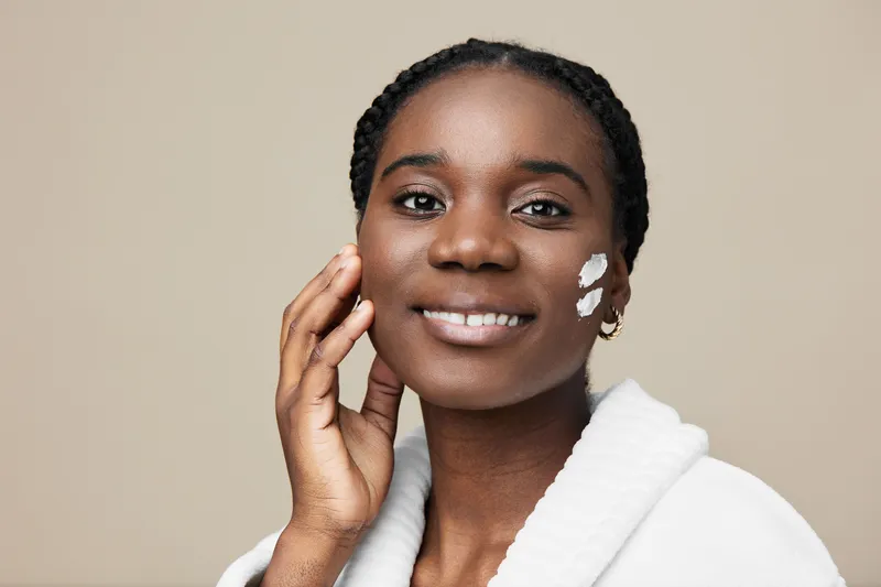 Young Black woman applying moisturizer on her face, smiling pleasantly against a soft beige background, conveying a sense of skincare beauty and self care