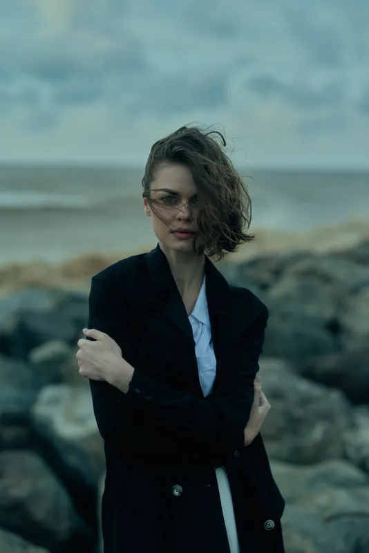 Woman in a dark coat stands on a rocky coast, wind tousling hair, by the sea, creating a moody editorial mood with strong fashion flair and atmospheric outdoor styling