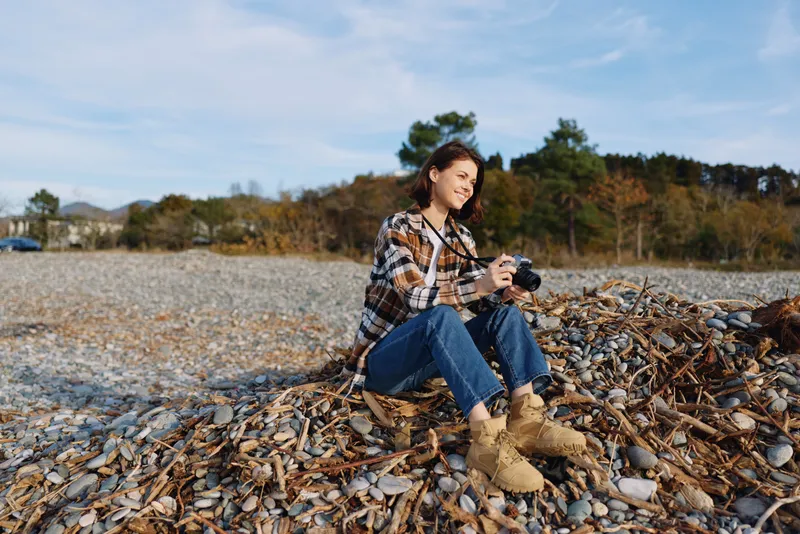 Woman photographer sitting on a rocky beach with a camera, autumn leaves around, casual outfit, capturing quiet landscape and nature scenes under a clear sky