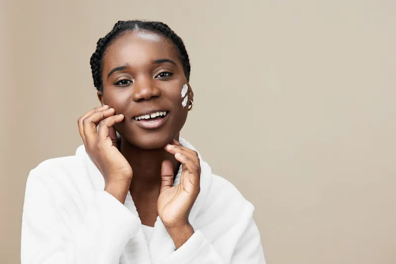 Smiling Black woman with natural skincare, wearing a white robe and facial cream, against a soft beige background, embodying wellness and beauty